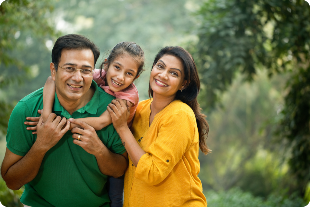 A couple standing in front of a wooded area, there's a little girl on the man's back with her arms around his neck